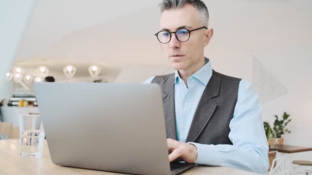 ガラスを身に着けている魅力的な大人の男は カフェの中に座っている間 彼のラップトップコンピュータを使用しています — ストック動画