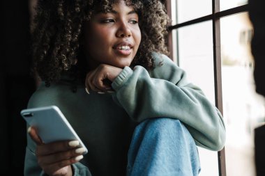 Pencere kenarında otururken cep telefonu kullanan Afro-Amerikan kadını.