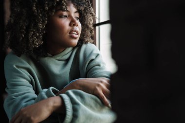 Odaklanmış Afro-Amerikalı kadın pencere eşiğinde otururken pencereye bakıyor.