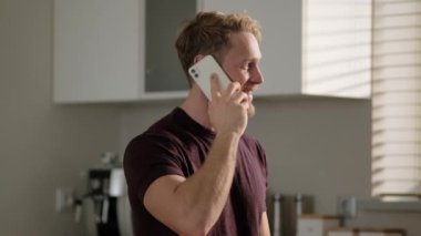 Gülümseyen sarışın bir adam akıllı telefonuyla konuşuyor. Evdeki mutfakta oturuyor.