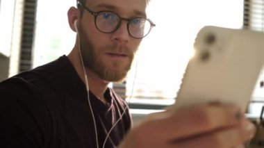 Gözlük takan ve kulaklık takan genç bir adam akıllı telefonunu evde oturuyor.