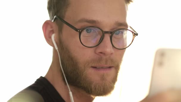一个戴着眼镜和耳机的积极的年轻人正坐在家里用他的智能手机 — 图库视频影像