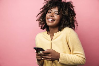 Komik Afrikalı Amerikalı kadın gülümsüyor ve pembe duvarda izole edilmiş cep telefonu kullanıyor.