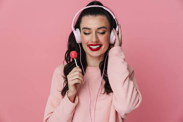Портрет веселой молодой девушки, стоящей в изоляции на розовом фоне, в наушниках, держащей леденец