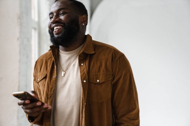 Neşeli Afrikalı Amerikalı adam gülümsüyor ve cep telefonunu kapalı alanda kullanıyor.