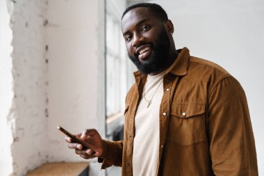 Neşeli Afrikalı Amerikalı adam gülümsüyor ve cep telefonunu kapalı alanda kullanıyor.