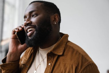 Neşeli Afrikalı Amerikalı adam gülümsüyor ve cep telefonuyla konuşuyor.