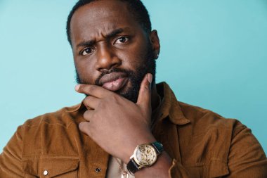 Memnun olmayan Afrikalı Amerikalı adam mavi arka planda izole edilmiş kameraya bakıyor ve düşünüyor.