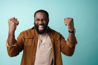 Heyecanlı Afrikalı Amerikalı adam, mavi arka planda izole bir şekilde zafer jesti yapıyor ve çığlık atıyor.