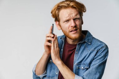 Kafası karışmış kızıl saçlı adam kameraya bakıyor ve beyaz arka planda izole edilmiş cep telefonu kullanıyor.
