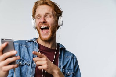 Kulaklıklı gülen kızıl saçlı adam beyaz arka planda izole edilmiş bir cep telefonu kullanıyor.
