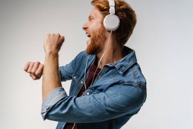Beyaz arka planda şarkı söyleyip dans eden kulaklıklı mutlu kızıl saçlı adam.