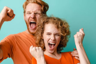 Heyecanlı kızıl saçlı genç bir çift çığlık atıyor ve kazanan jestler yapıyor mavi arka planda izole edilmiş