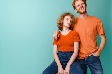 Genç, mutlu, kızıl saçlı, gülümseyen ve kameraya sarılan çift mavi arka planda izole edilmiş.