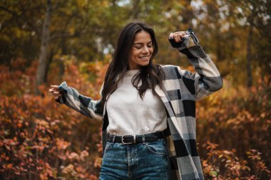 Güzel esmer mutlu kadın sonbahar ormanlarında gezinirken dans ediyor.