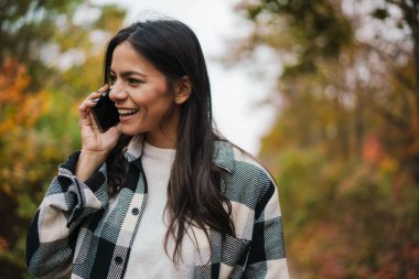 Güzel esmer, mutlu kadın sonbahar ormanlarında gezinirken cep telefonuyla konuşuyor.