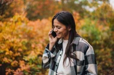 Gülümseyen çekici kadın sonbahar parkında yürürken cep telefonu kullanıyor.