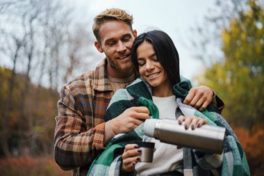 Genç mutlu çift sonbahar ormanlarında piknik yaparken çay içip sarılıyorlar.