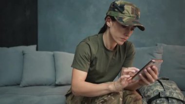 A concentrated young woman wearing military uniform is using her smartphone while sitting on the sofa inside the house