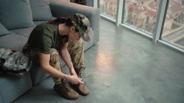A beautiful young woman wearing military uniform is putting on soldier boots while sitting inside the house before leaving