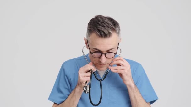 Zadowolony Starszy Lekarz Mundurze Medycznym Zakładający Stetoskop Stojący Szarym Tle — Wideo stockowe