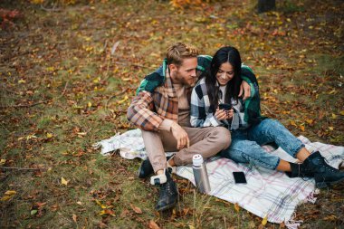 Genç mutlu çift sonbahar ormanında dinlenirken çay içip sarılıyorlar.