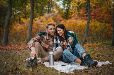 Genç mutlu çift sonbahar ormanında dinlenirken çay içip sarılıyorlar.