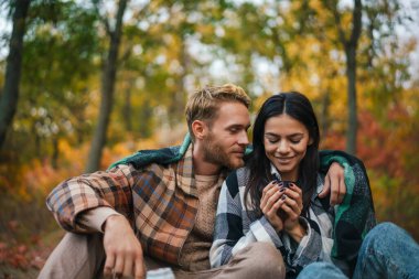 Genç mutlu çift sonbahar ormanında dinlenirken çay içip sarılıyorlar.