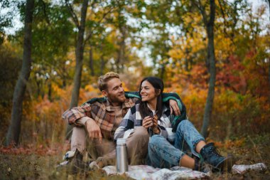 Genç mutlu çift sonbahar ormanında dinlenirken çay içip sarılıyorlar.