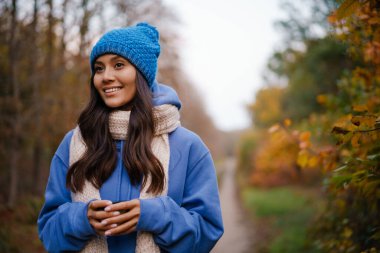 Güzel esmer, mutlu, örgü şapkalı, sonbahar ormanlarında gezinirken çay içen bir kadın.
