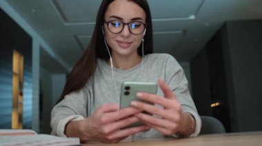 A pleased woman with earphones is using her smartphone sitting inside the apartments
