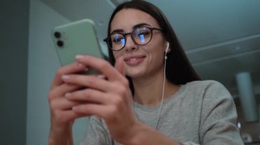 A smiling woman wearing glasses is using her smartphone sitting inside the apartments