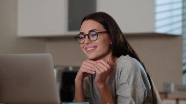 A smiling woman is talking with video connection using her laptop sitting inside the apartments