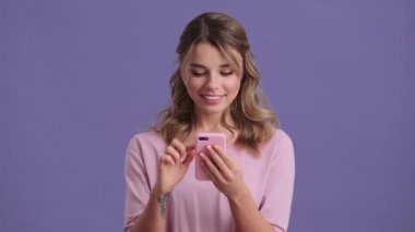 A surprised woman is using her smartphone standing isolated over a violet background