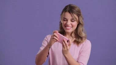 A pleased woman is playing games using her smartphone standing isolated over a violet background