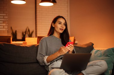 Güzel bir kadının evdeki kanepede dizüstü bilgisayarla çalışırken cep telefonu kullanması.