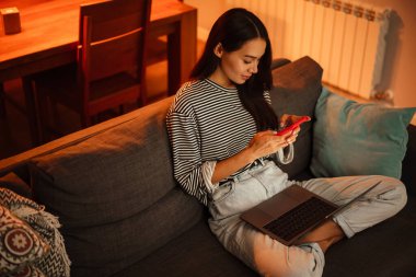 Evde kanepede dizüstü bilgisayarla çalışırken cep telefonu kullanan güzel bir kadın.