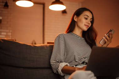 Evde kanepede dizüstü bilgisayarla çalışırken cep telefonu kullanan güzel bir kadın.