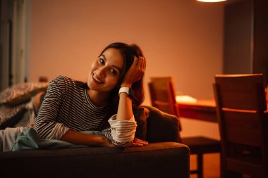 Evdeki kanepede otururken kameraya gülümseyen neşeli güzel kadın.