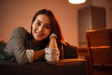 Evdeki kanepede otururken kameraya gülümseyen neşeli güzel kadın.