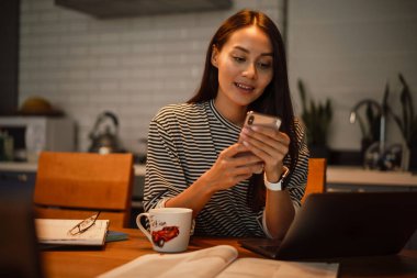 Ev mutfağında dizüstü bilgisayarla çalışırken cep telefonu kullanan sevimli bir kadın.