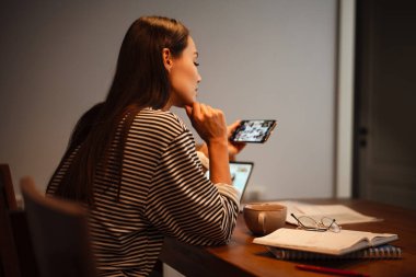 Ev mutfağında dizüstü bilgisayarla çalışırken cep telefonu kullanan çekici bir kadın.