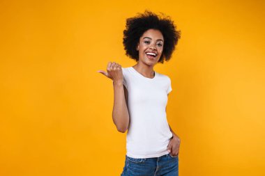 Mutlu Afrikalı Amerikalı kız gülümsüyor ve sarı arka planda izole edilmiş kopyalama alanını işaret ediyor.