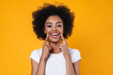 Mutlu Afrikalı Amerikalı kız sarı arka planda izole edilmiş gülümsemesini işaret ediyor.