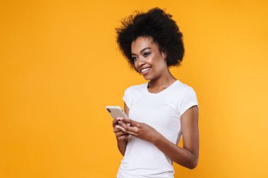 Mutlu, genç, Afro-Amerikan kadın gülümseyen ve elinde cep telefonu tutan sarı arka planda.