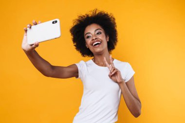 Mutlu Afrikalı genç Amerikalı kadın sarı arka planda izole edilmiş cep telefonundan selfie çekerken zafer işareti gösteriyor.