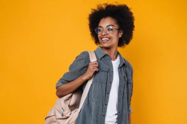 Gözlüklü neşeli Afrikalı Amerikalı kız sarı arka planda izole edilmiş kameraya bakıp gülüyor.