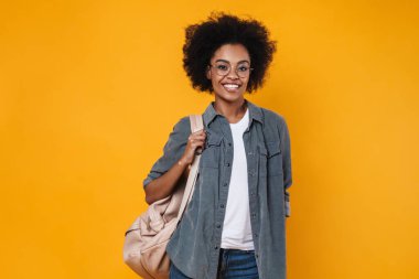 Gözlüklü neşeli Afrikalı Amerikalı kız sarı arka planda izole edilmiş kameraya bakıp gülüyor.