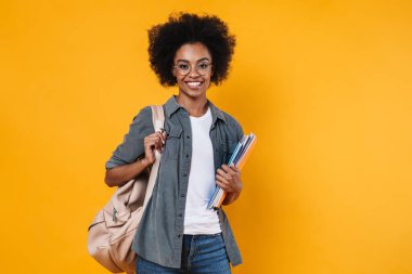 Gözlüklü neşeli Afrikalı Amerikalı kız sarı arka planda izole edilmiş egzersiz kitaplarıyla poz veriyor.