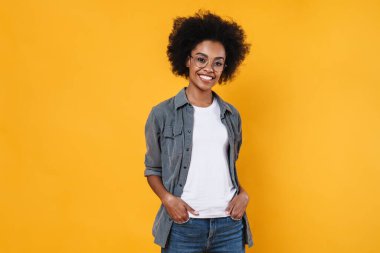 Gözlüklü neşeli Afrikalı Amerikalı kız sarı arka planda izole edilmiş kameraya bakıp gülümsüyor.